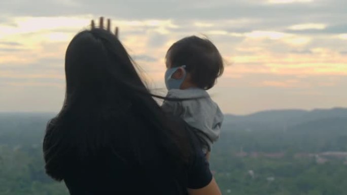 日落时快乐家庭的后视图，亚洲母亲和男婴儿子年龄17个月戴着防护口罩，自由地看着山上的自然景色，教孩子