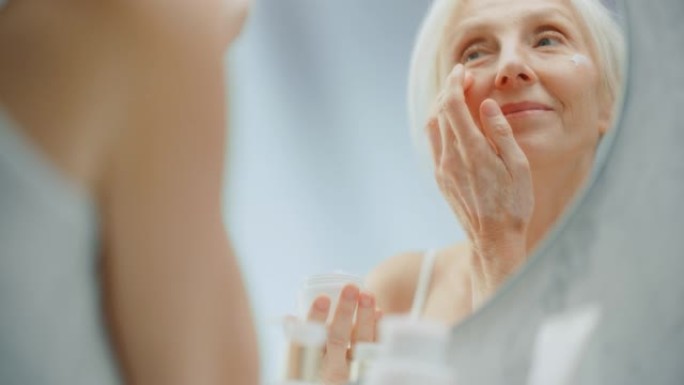 美丽的高级女人的肖像早晨例行，照镜子轻轻涂抹面霜。老年女士用天然抗衰老化妆品，产品使皮肤柔软，光滑，
