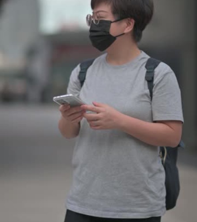 亚洲华人中年妇女戴着防护面罩，白天在城市街道上使用智能手机