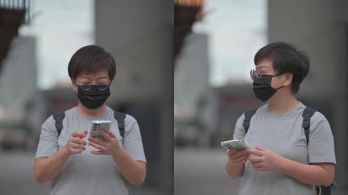 亚洲华人中年妇女戴着防护面罩，白天在城市街道上使用智能手机