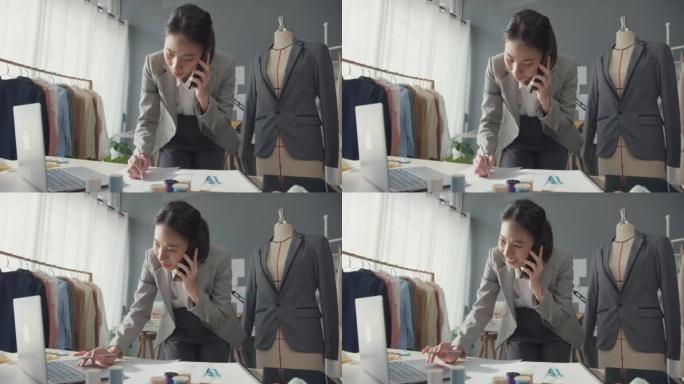 专业开朗的亚洲女商人设计师坐在办公桌上使用笔记本电脑呼叫智能手机，以在工作室工作场所在线市场上确认客