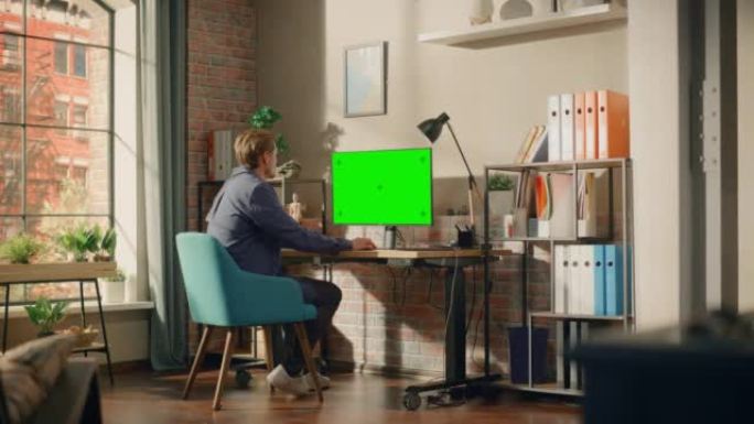 年轻英俊的男人在家工作，在带有绿屏模拟显示的台式计算机上。创意男性查看社交媒体，浏览互联网。明亮阁楼