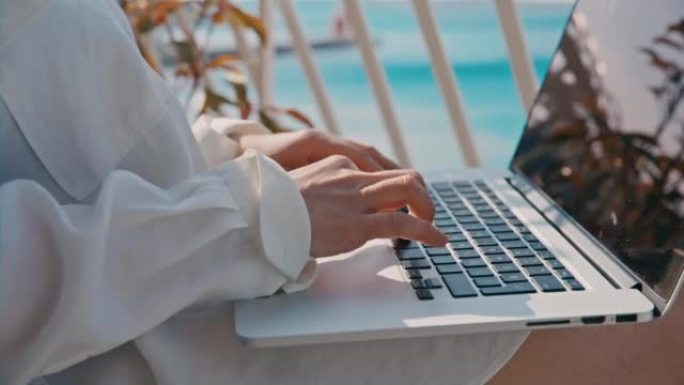特写女人在笔记本电脑键盘上打字，坐在酒店房间的阳台上，可以看到大海