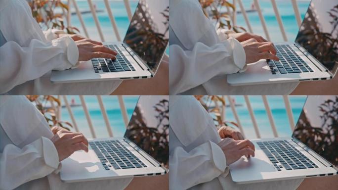 特写女人在笔记本电脑键盘上打字，坐在酒店房间的阳台上，可以看到大海