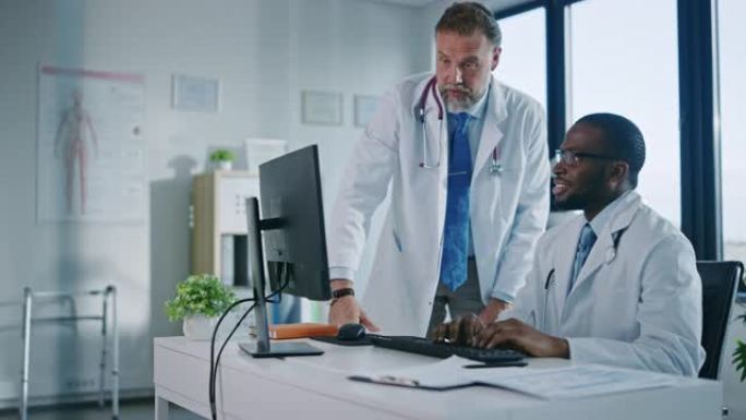 年轻的非洲裔美国医生正在一家医疗诊所与一名高级医生交谈。同事在医院办公室讨论台式计算机上的工作。助理