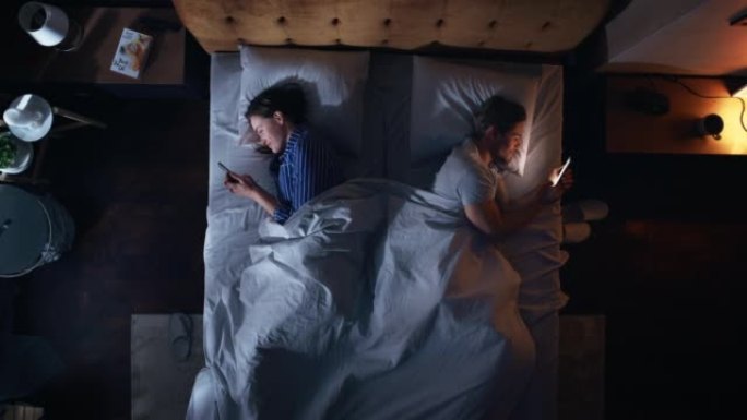 顶视图公寓卧室: 白人夫妇躺在床上，都使用智能手机。两口之家使用手机浏览社交媒体，交流，搜索互联网，