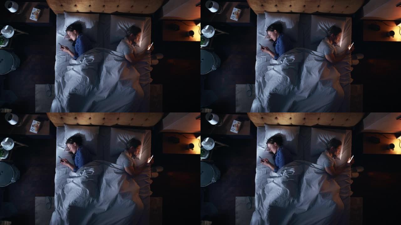 顶视图公寓卧室: 白人夫妇躺在床上，都使用智能手机。两口之家使用手机浏览社交媒体，交流，搜索互联网，