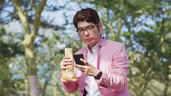 亚洲男子坐在公园里吃零食并使用智能手机