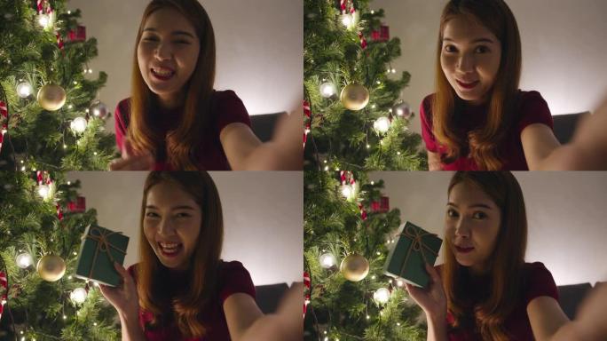 年轻的亚洲女性使用智能手机视频通话与圣诞礼物盒子的情侣交谈，圣诞树装饰着家里客厅的装饰品。