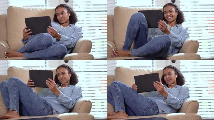 使用平板电脑，耳塞坐在扶手椅上的年轻混血妇女