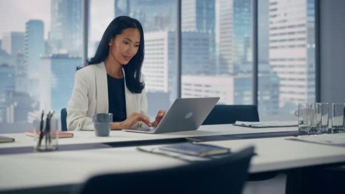 在大城市办公室，穿着时髦西装在笔记本电脑上工作的成功亚洲女商人的肖像。强大的CEO管理数字电子商务项
