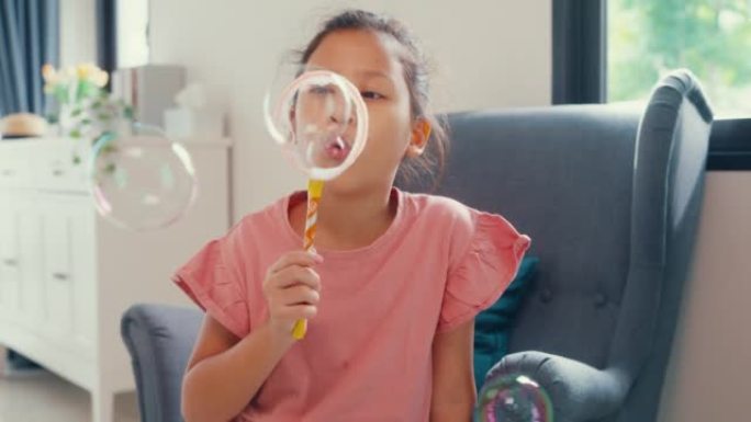 快乐的亚洲蹒跚学步的小女孩坐在沙发椅上玩泡泡在镜头前带着自然的微笑在家里的客厅玩得开心快乐。