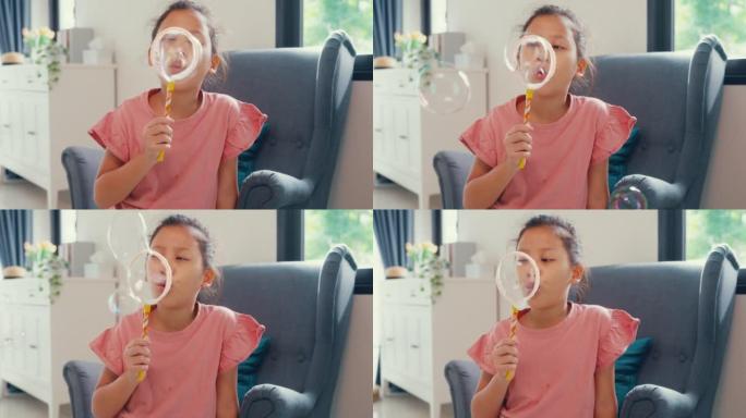 快乐的亚洲蹒跚学步的小女孩坐在沙发椅上玩泡泡在镜头前带着自然的微笑在家里的客厅玩得开心快乐。