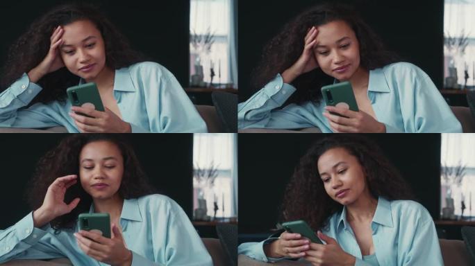 快乐的年轻有吸引力的轻松的非洲自由职业者商业女人微笑着使用智能手机应用程序在家沙发上工作。
