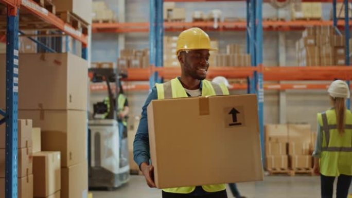 帅气的男工人戴着安全帽，拿着纸板箱，穿过摆满货架的零售仓库。在物流配送中心工作。前跟随慢动作