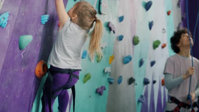 小孩在运动中心爬人造墙，女人在背景中使用安全设备