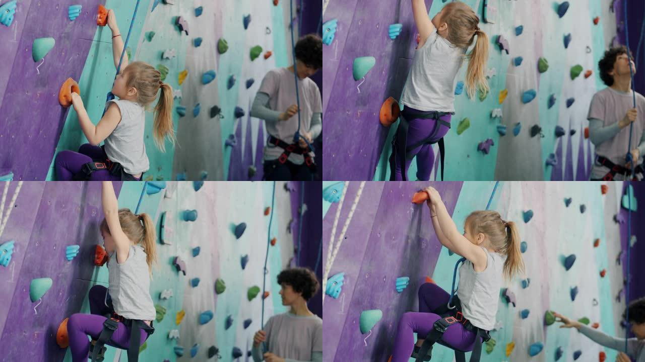 小孩在运动中心爬人造墙，女人在背景中使用安全设备