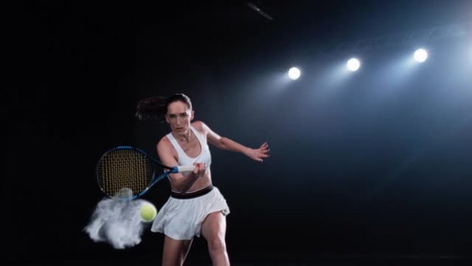 一名黑色背景的运动女网球运动员在聚光灯下击中一个低球的美学镜头。电影般的超级慢动作捕捉到了一个具有烟
