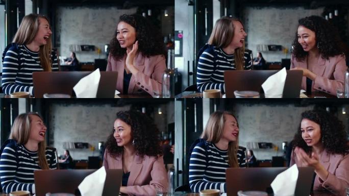 在现代咖啡店会议上，两个美丽的快乐微笑的年轻多民族朋友妇女交谈，大笑和使用笔记本电脑。