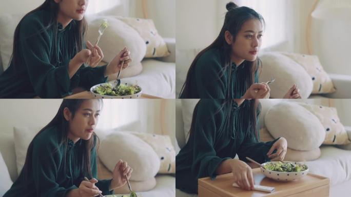 年轻快乐的亚洲女人坐在客厅的沙发上吃健康的沙拉。
