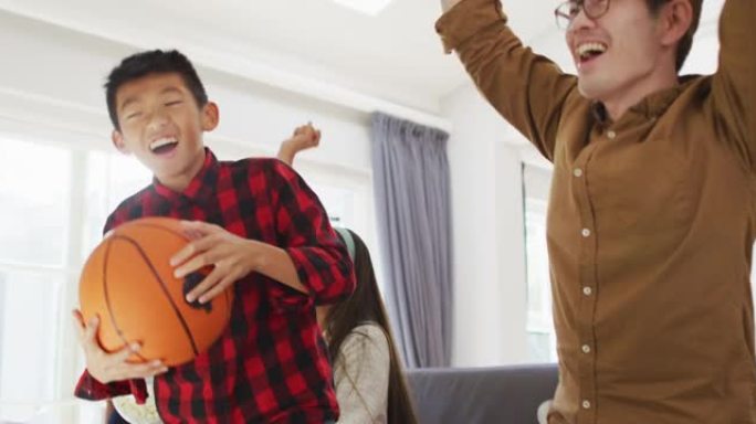 激动的亚洲父母带着女儿和儿子坐在沙发上，在电视上看篮球，欢呼