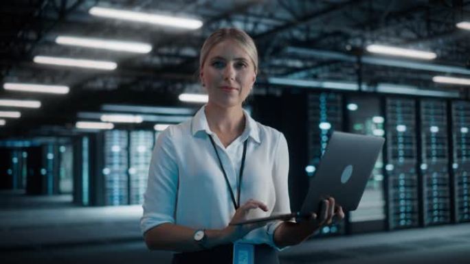 成功的女性首席技术官的肖像使用笔记本电脑，站在大仓库数据中心，看着相机。SAAS、云服务服务器的系统