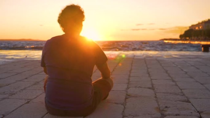 低角度: 年轻人坐在石头地上，在日落时观察大海。