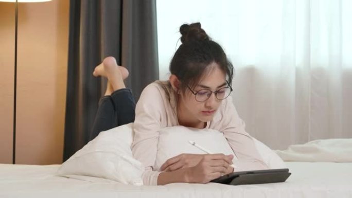 一个年轻美丽的眼镜亚洲妇女躺在床上，使用平板电脑在网上工作或学习的POV。检疫期间在家穿着便服的漂亮