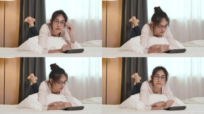 一个年轻美丽的眼镜亚洲妇女躺在床上，使用平板电脑在网上工作或学习的POV。检疫期间在家穿着便服的漂亮