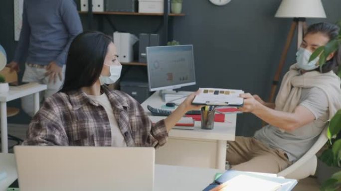 戴着医用口罩的年轻女子在办公室工作，使用笔记本电脑并与同事合作