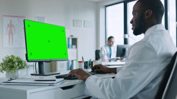 非裔美国医生正在健康诊所使用带有绿屏模拟显示的计算机。穿着白大褂的医生正在医院办公室的桌子后面阅读病