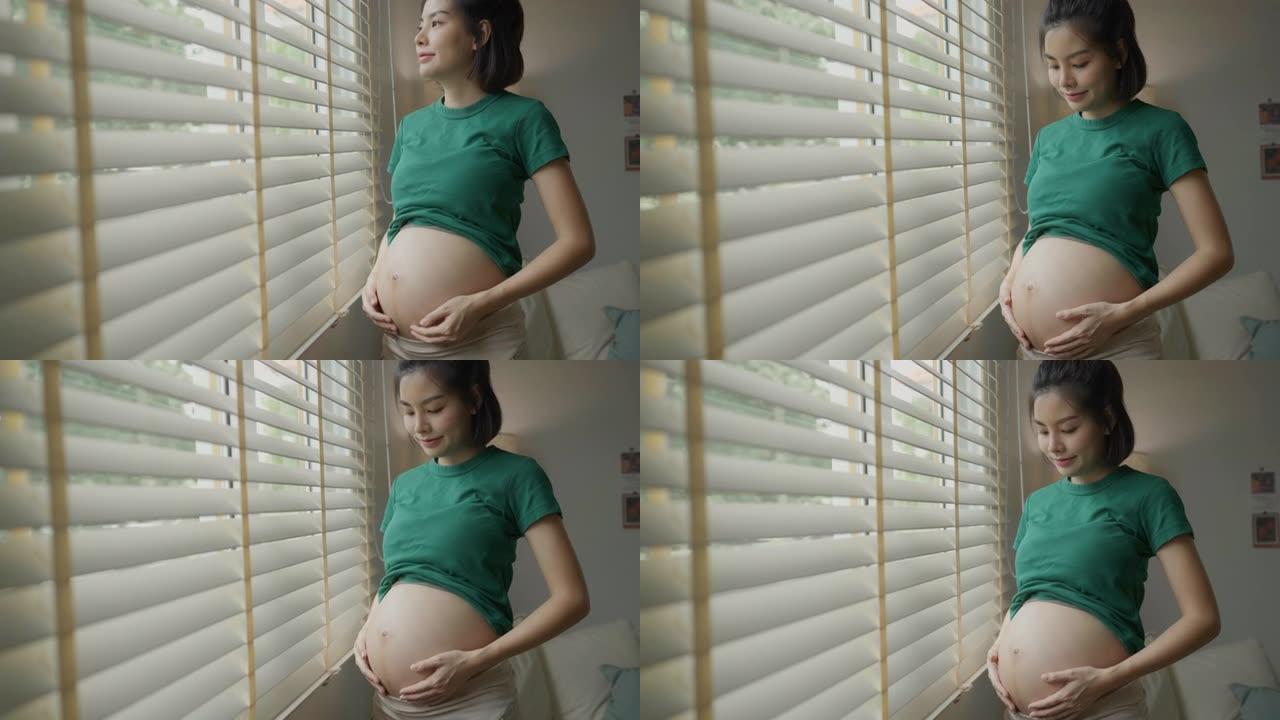 一位年轻的孕妇用手靠近窗户抚摸腹部。