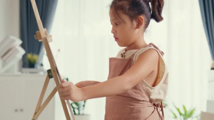 特写带围裙的亚洲学步女孩坐在沙发椅上，在带调色板的油画画布前使用画笔专注于周末在家绘画抽象。