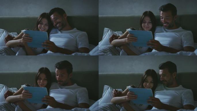 年轻快乐微笑的真实照片刚刚结婚的情侣在一起享受时光，同时使用平板电脑进行家庭娱乐，晚上在家里卧室的床