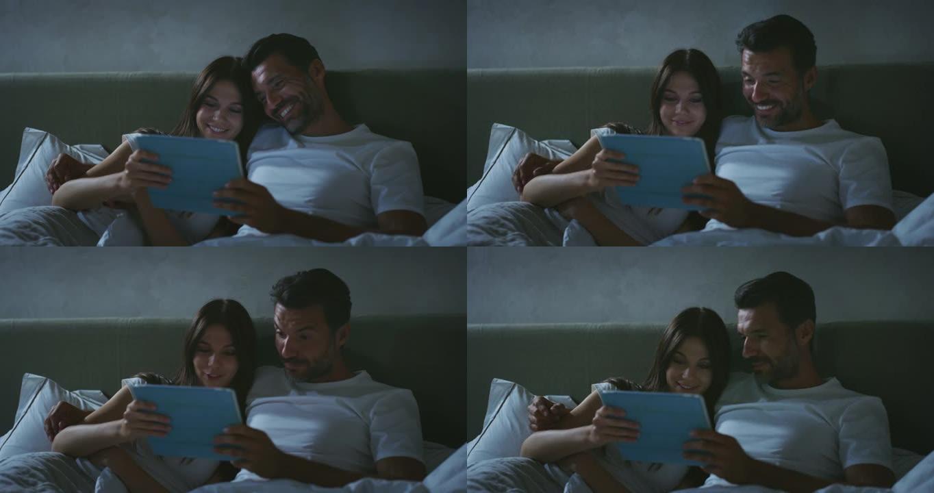 年轻快乐微笑的真实照片刚刚结婚的情侣在一起享受时光，同时使用平板电脑进行家庭娱乐，晚上在家里卧室的床