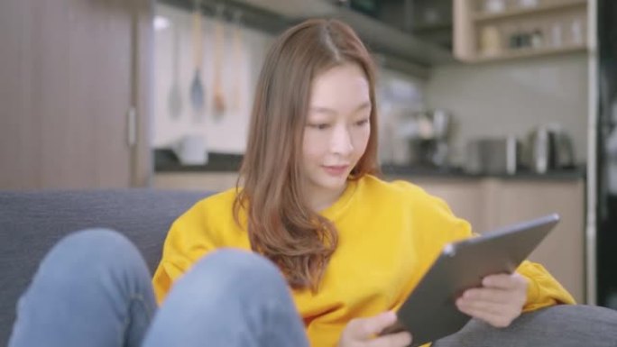 美丽的亚洲年轻女子，年龄36岁，穿着彩色黄色衬衫，在家工作，通过数字平板电脑远程团队合作，早上在家坐