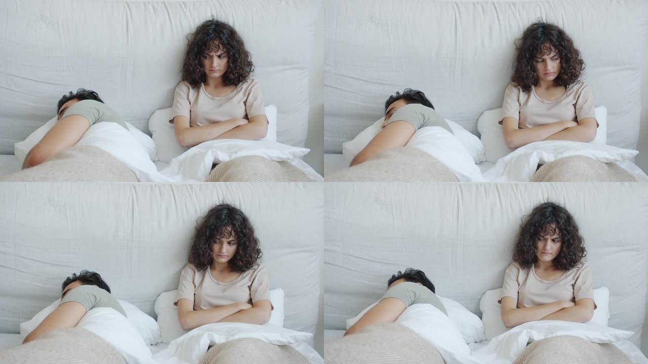 愤怒的白人妇女看着躺在床上的熟睡的丈夫阿拉伯男人