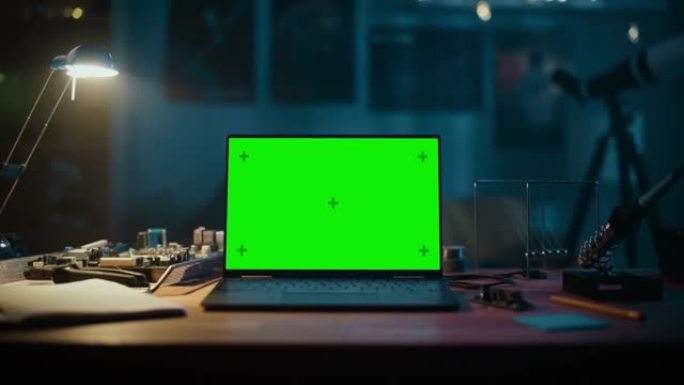 笔记本电脑站在一张绿屏Chromakey模拟显示器的桌子上。舒适的空房间，桌子上有灯，电路板，牛顿的
