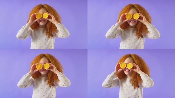 在紫色工作室背景下，红发年轻女孩像眼镜一样在脸前拿着两半橘子的工作室镜头-慢动作拍摄