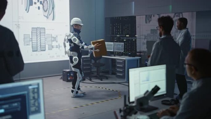在开发实验室中: 工程师和科学家看着机器人外骨骼演示，有人举起沉重的Carboard Box，人们在