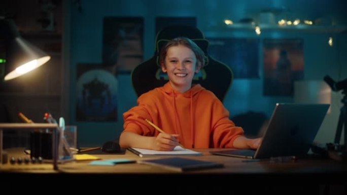 一个穿着橙色运动衫的聪明小男孩在家里舒适的黑暗房间里的笔记本电脑上做作业的肖像。少年快乐，看着镜头微