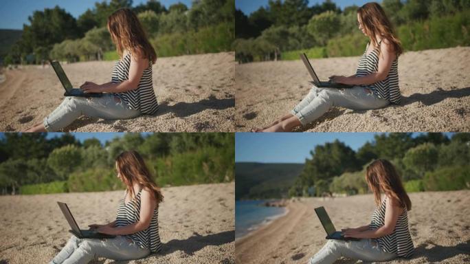 年轻女子坐在沙滩上，在海滩上的笔记本电脑上工作