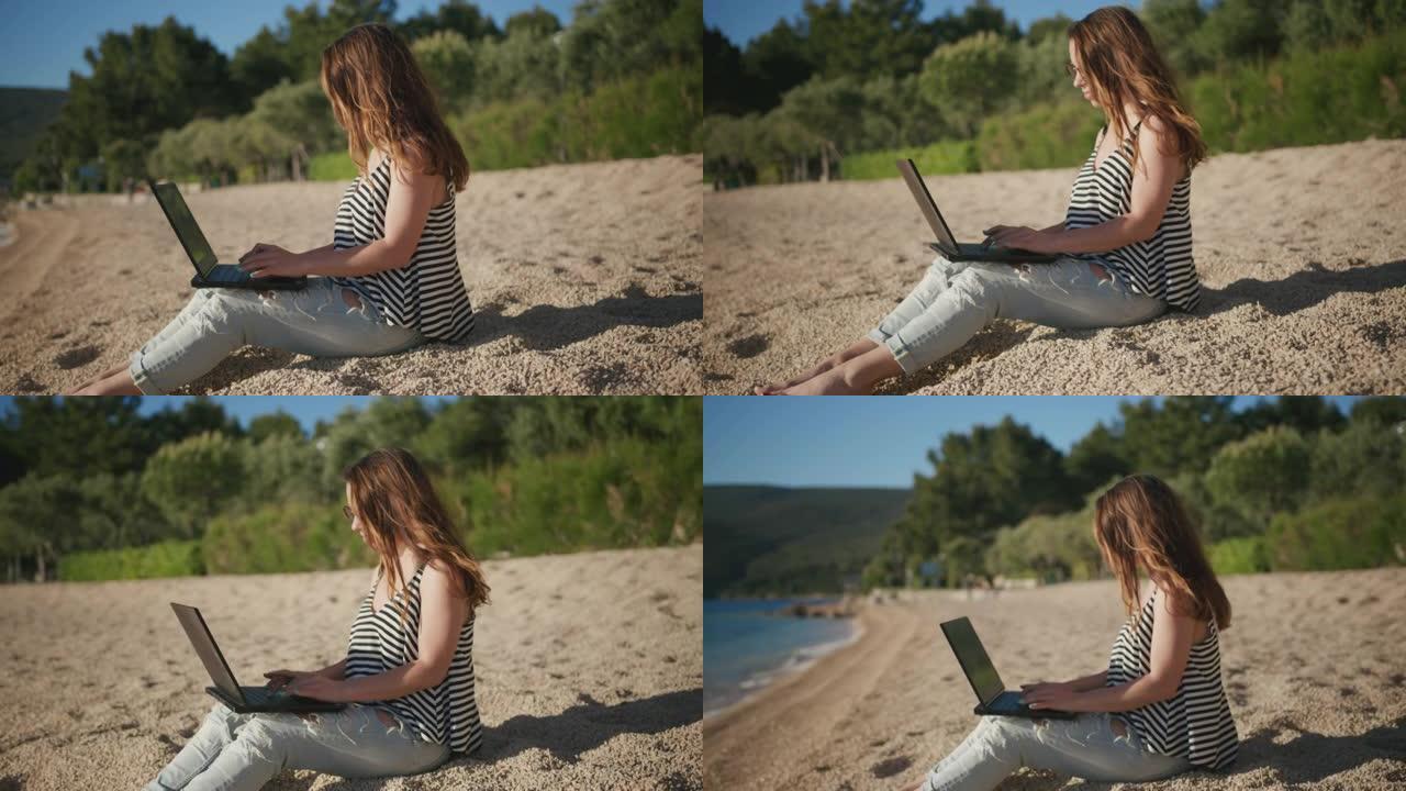 年轻女子坐在沙滩上，在海滩上的笔记本电脑上工作