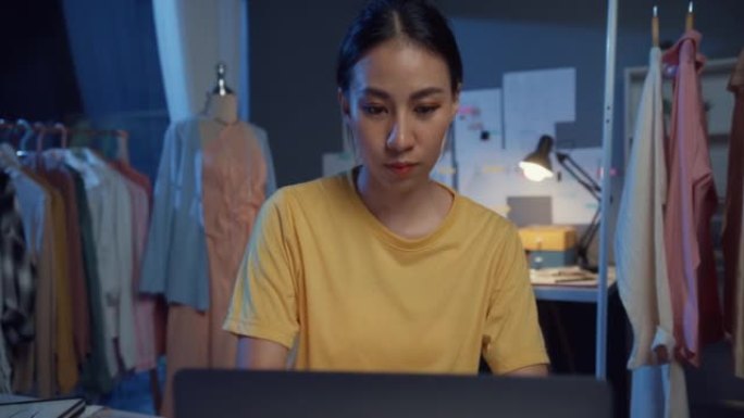 专业亚洲女性时装设计师，休闲使用笔记本电脑平面设计服装思维创意，在夜间创意办公室创造新系列。裁缝裁缝