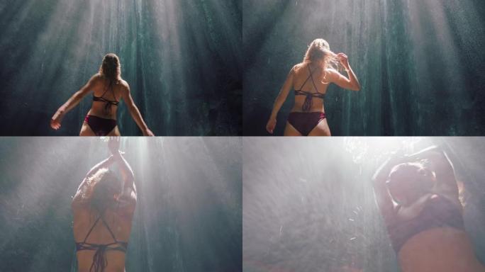 美丽的女人穿着比基尼在瀑布洞里跳舞，享受双臂举起4k的自由