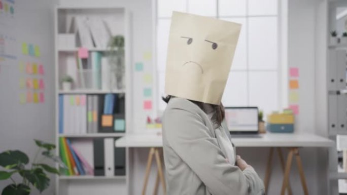 年轻的亚洲女商人坐在办公桌上，穿着悲伤的面具纸工艺包，在办公室工作失败时心情不好。