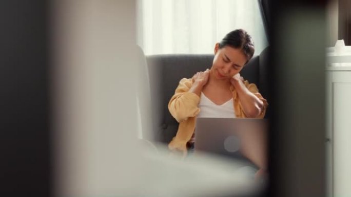 年轻的亚洲女女孩或大学生坐在沙发椅子上，用电脑笔记本电脑感到疼痛，在家里的客厅因加班而受伤。