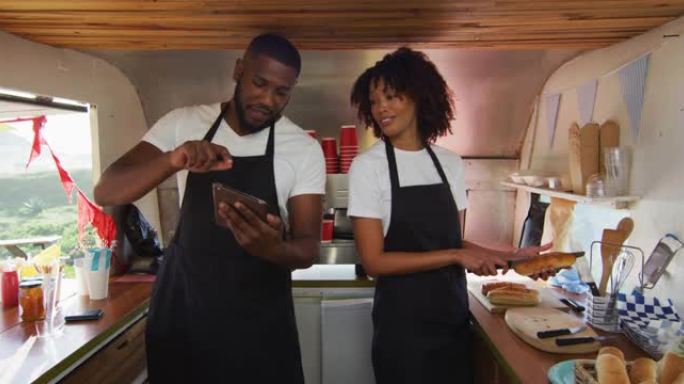 非裔美国夫妇穿着使用数字平板电脑的围裙，并在餐车中准备热狗
