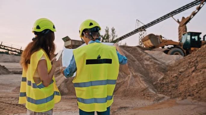 两名采矿工程师的慢动作，穿着全套防护装备，在采石场前检查大型蓝图图