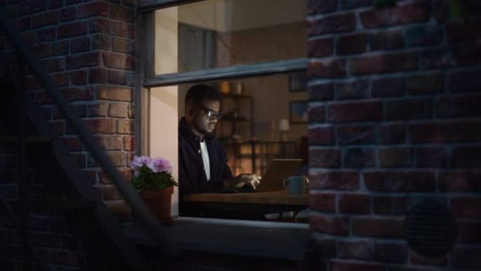 夜间在舒适温暖的公寓里，在家里用笔记本电脑工作的黑人创意年轻人的肖像。非裔美国男性开发人员工程软件。